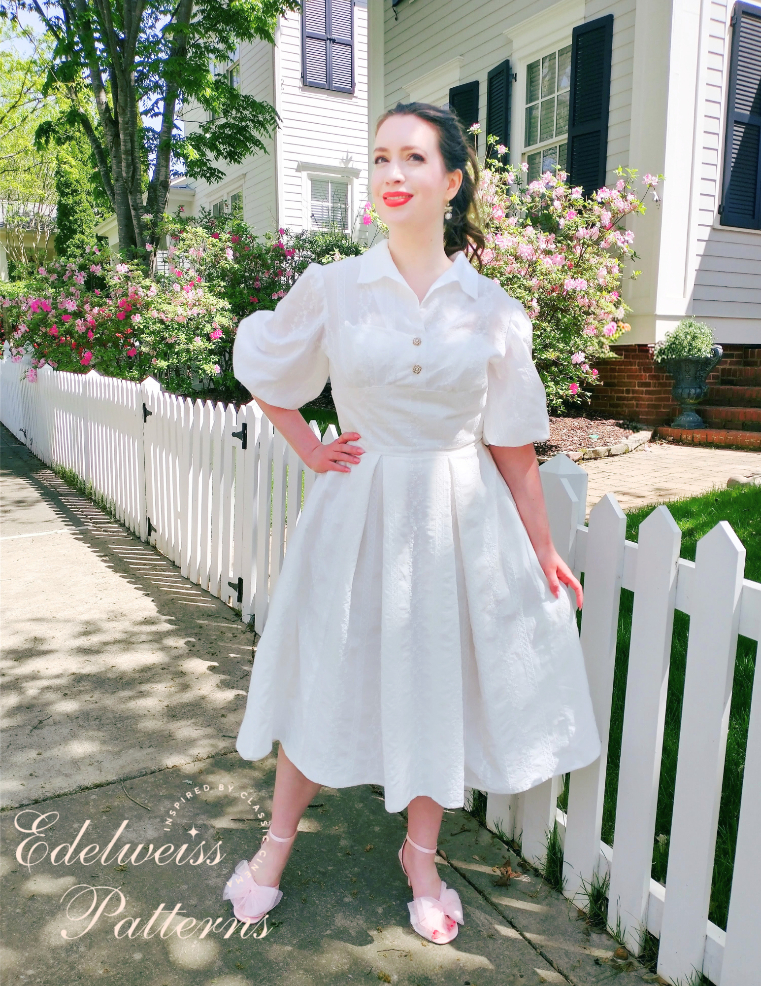 Lady wearing a vintage 1950s white shirt dress in front of a white picket fence in a beautiful neighborhood